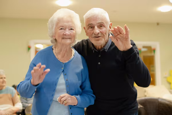 An elderly husband and wife standing arm in arm, smiling and waving together