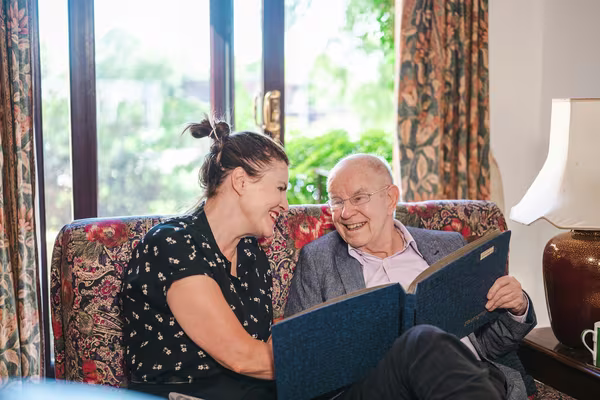 An elderly father and his adult daughter sitting together on a sofa, smiling as they look through a photo album
