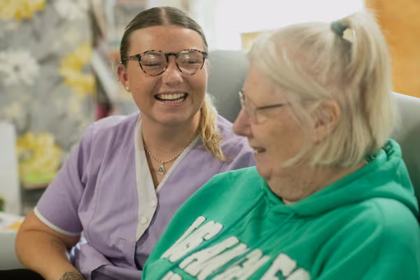 A smiling home care worker in a lilac uniform sitting and laughing with an older woman at home