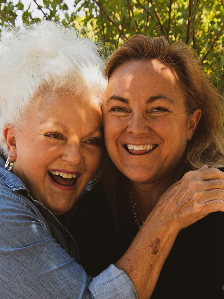 An older woman and her adult daughter embracing cheek to cheek outdoors, both laughing warmly