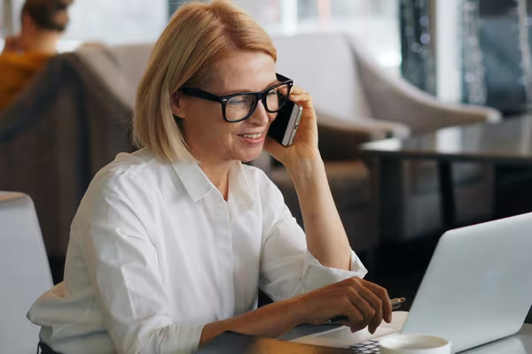 A woman wearing glasses on the phone while using a laptop at a table