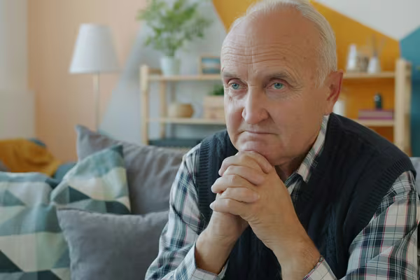 An older man sitting alone on his sofa at home, resting his chin on his clasped hands and looking thoughtful
