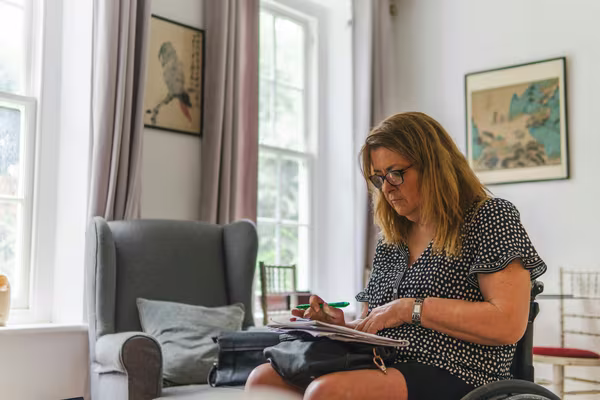 A woman sitting at home with a pen, working through paperwork and documents on her lap