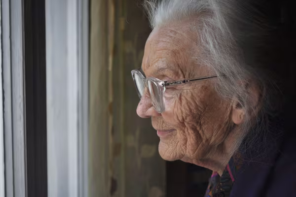 An older woman wearing glasses looking out of a window, deep in thought