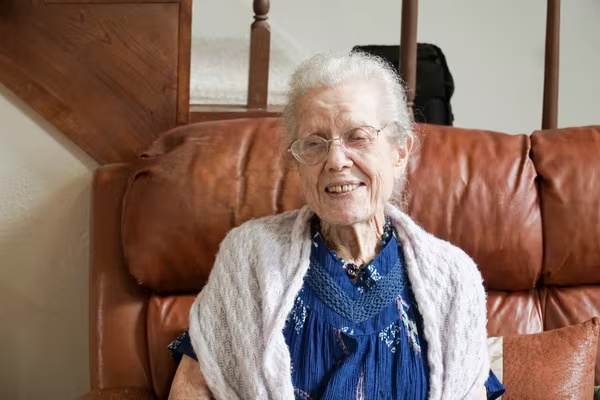 An older woman sitting smiling in an armchair at home, with a staircase behind her