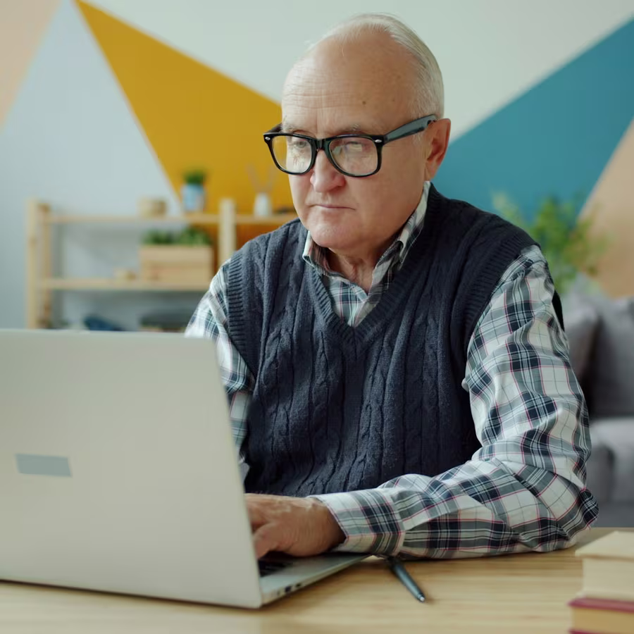 An older man wearing glasses sitting at a table at home, using a laptop
