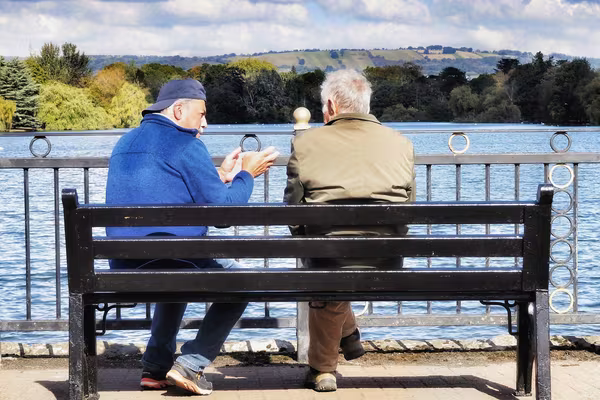 Two older men sitting together on a bench by a lake, talking