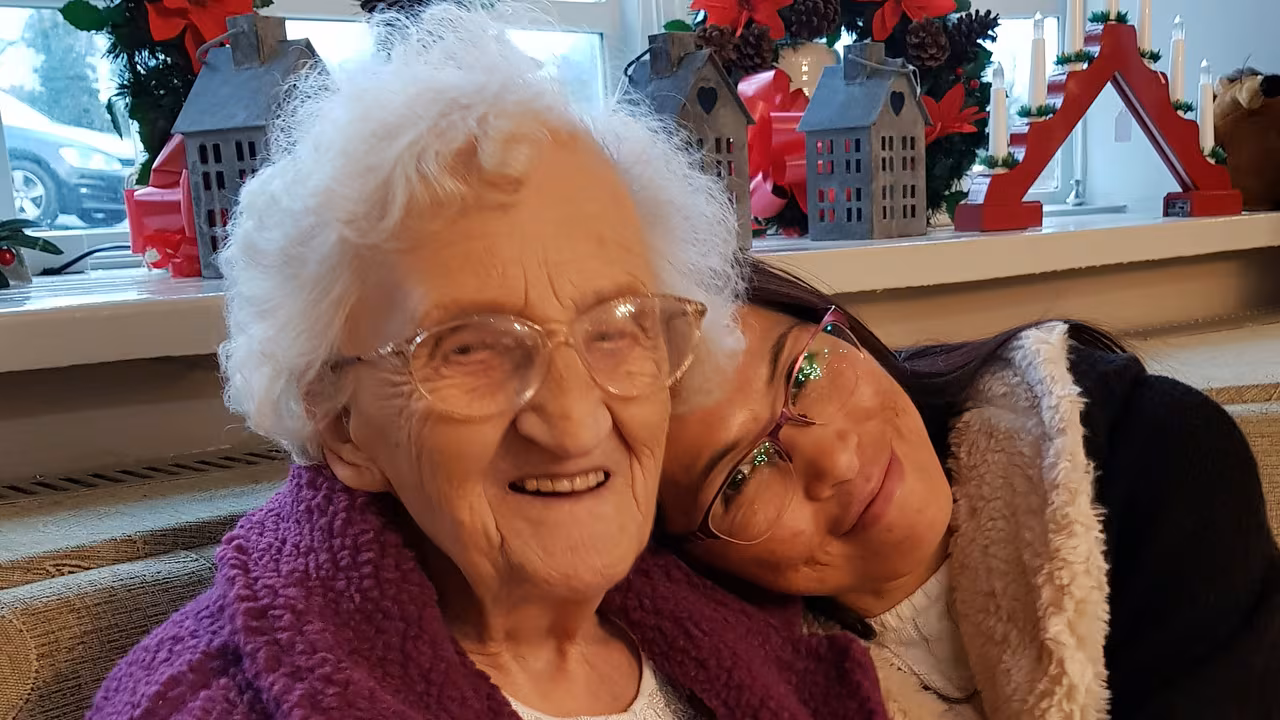 An older woman and a younger woman sitting cheek to cheek on a sofa, both smiling, with festive decorations behind them