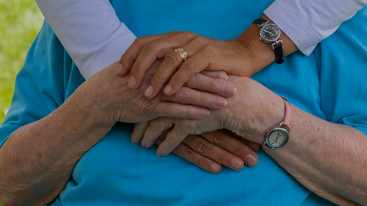 A younger person's arms wrapped gently around an older loved one, their hands clasped together