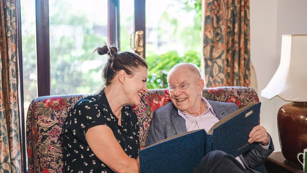 An elderly father and his adult daughter sitting together on a sofa, smiling as they look through a photo album