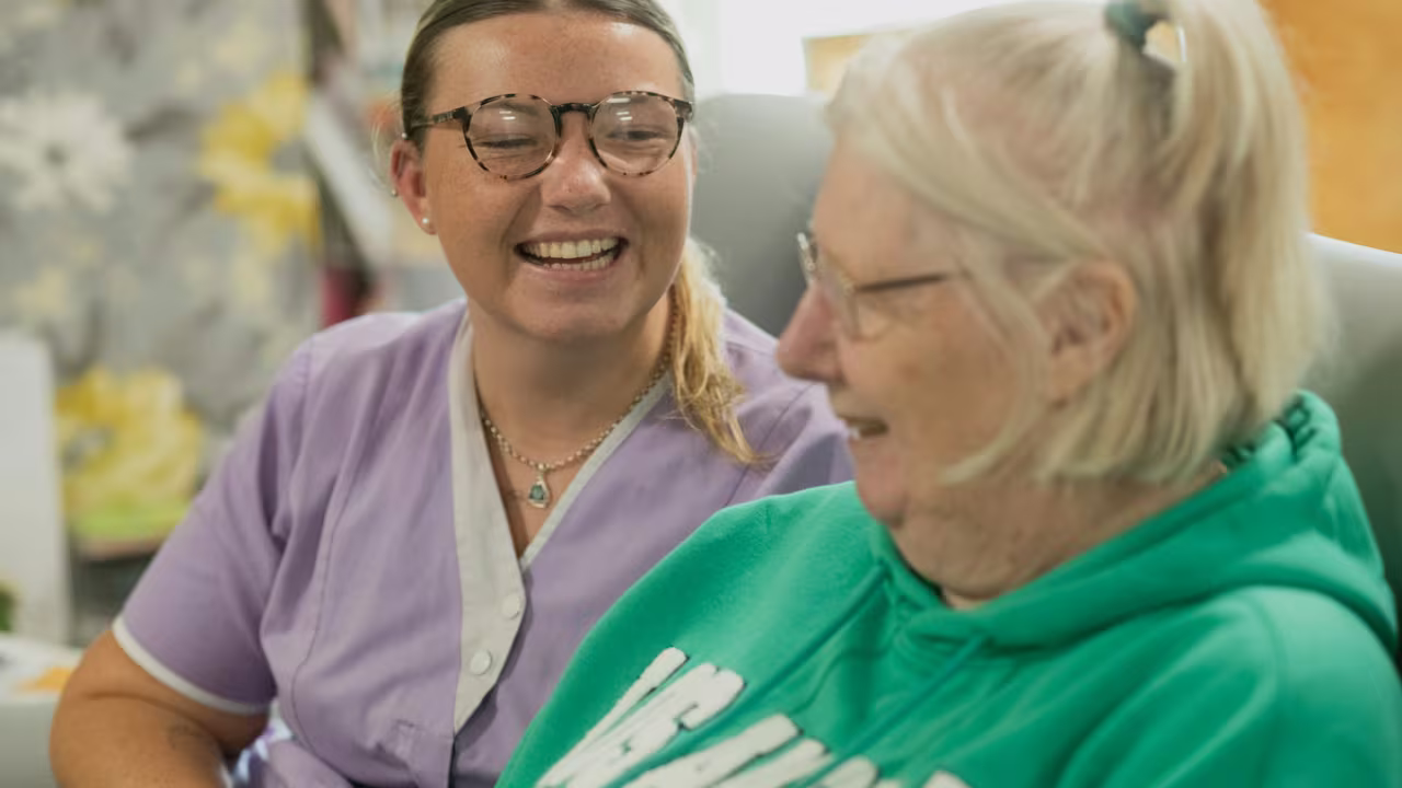 A smiling home care worker in a lilac uniform sitting and laughing with an older woman at home