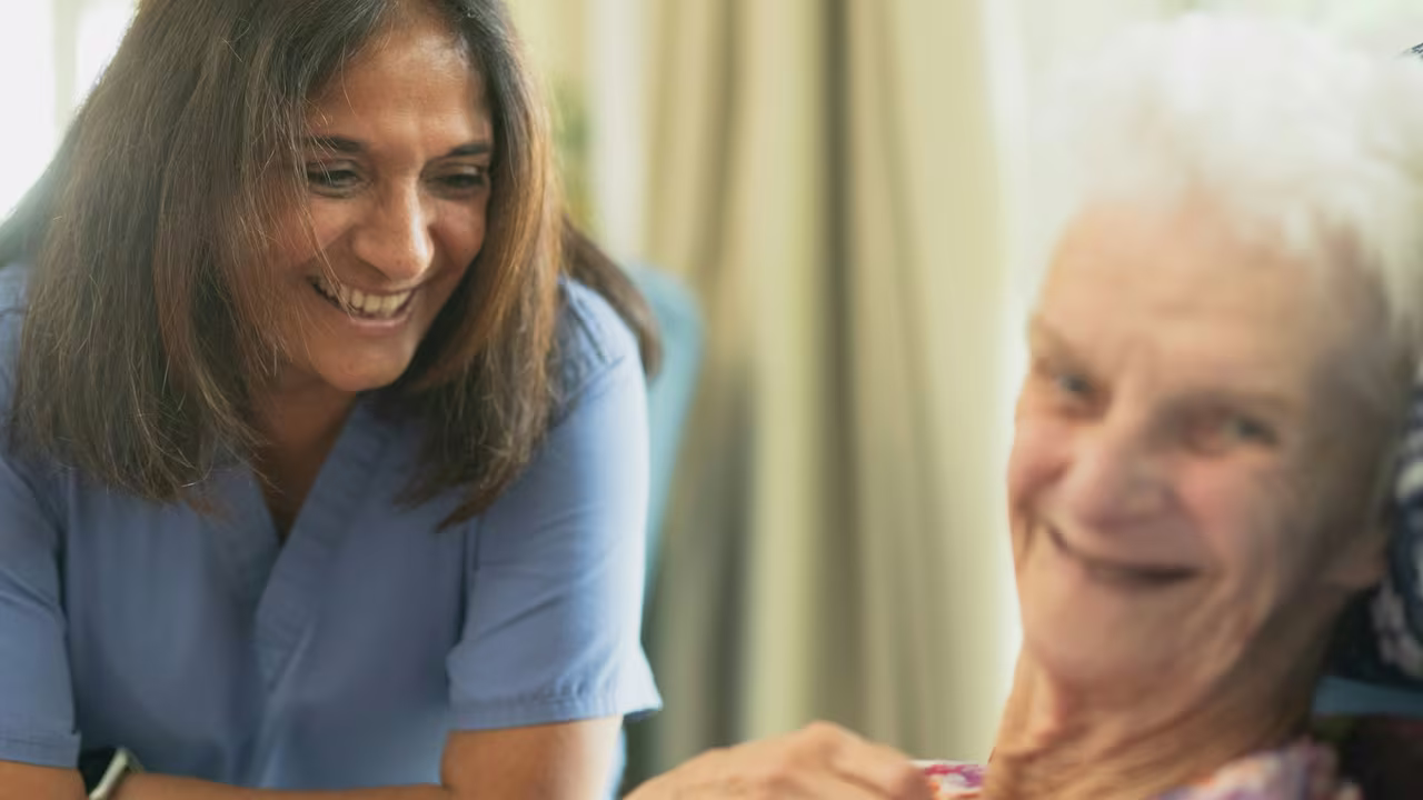 A smiling care worker in blue scrubs sharing a warm moment with an older woman at home