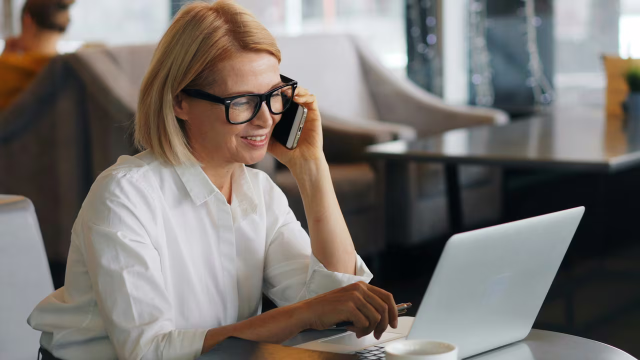A woman wearing glasses on the phone while using a laptop at a table