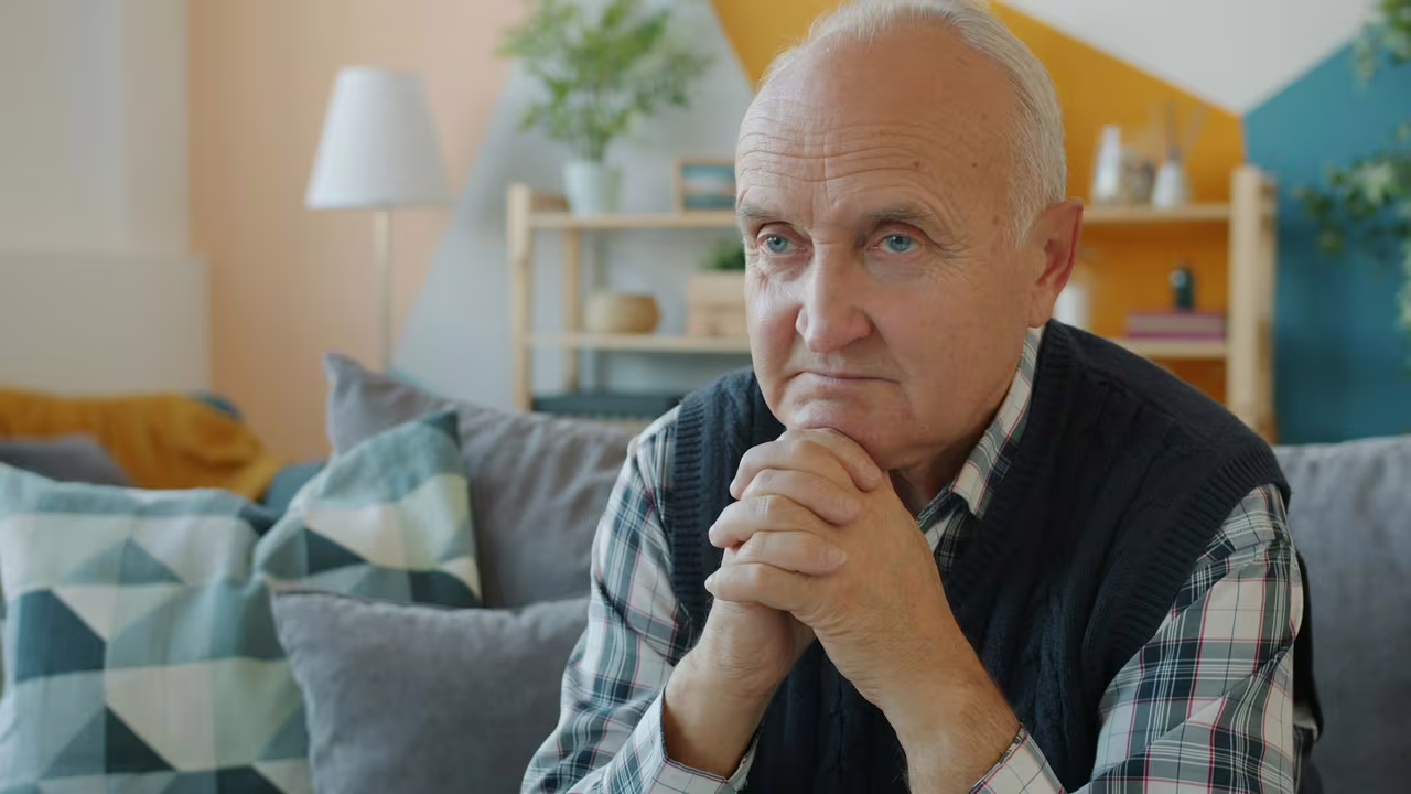 An older man sitting alone on his sofa at home, resting his chin on his clasped hands and looking thoughtful