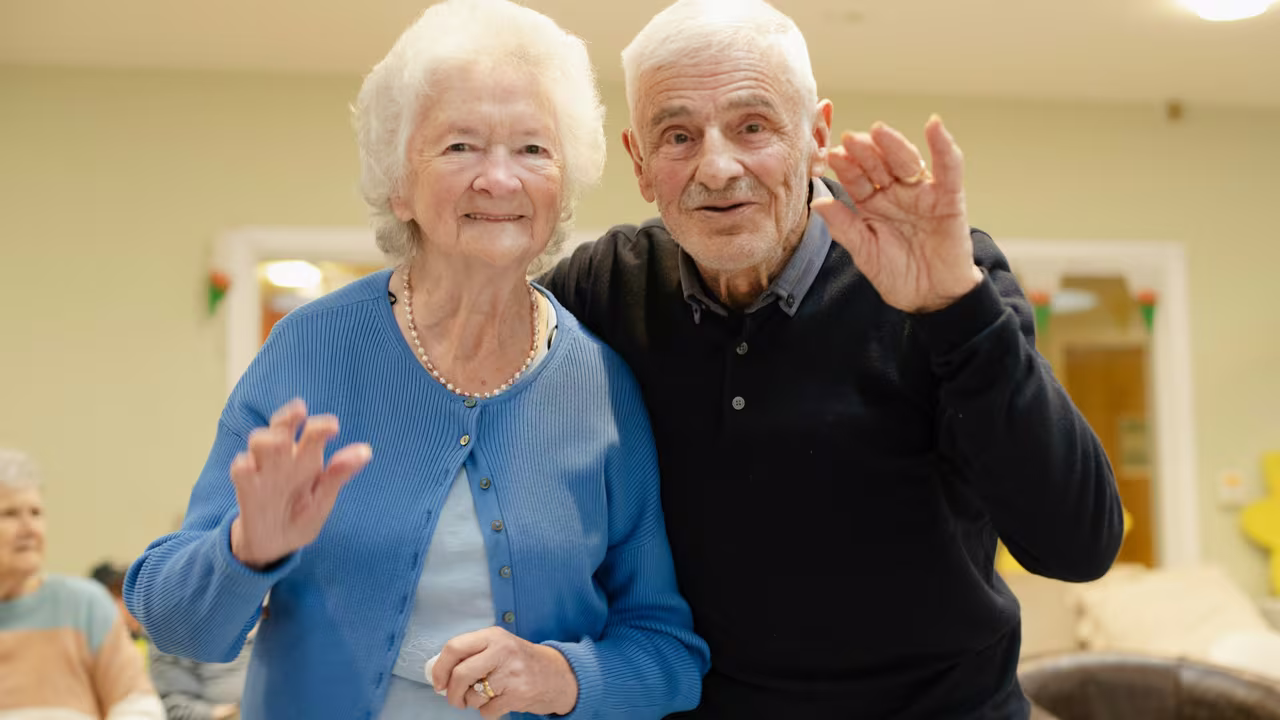 An elderly husband and wife standing arm in arm, smiling and waving together