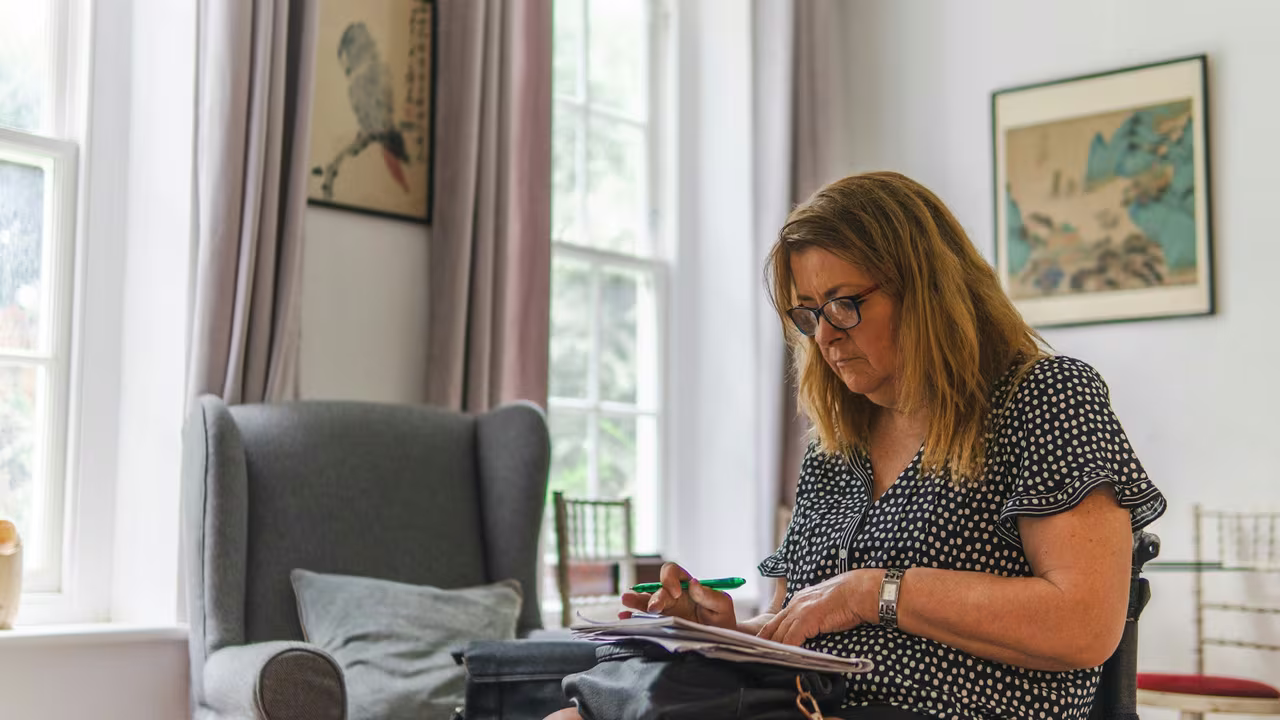 A woman sitting at home with a pen, working through paperwork and documents on her lap