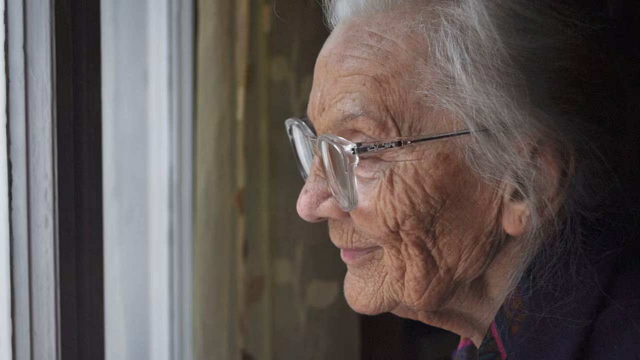 An older woman wearing glasses looking out of a window, deep in thought