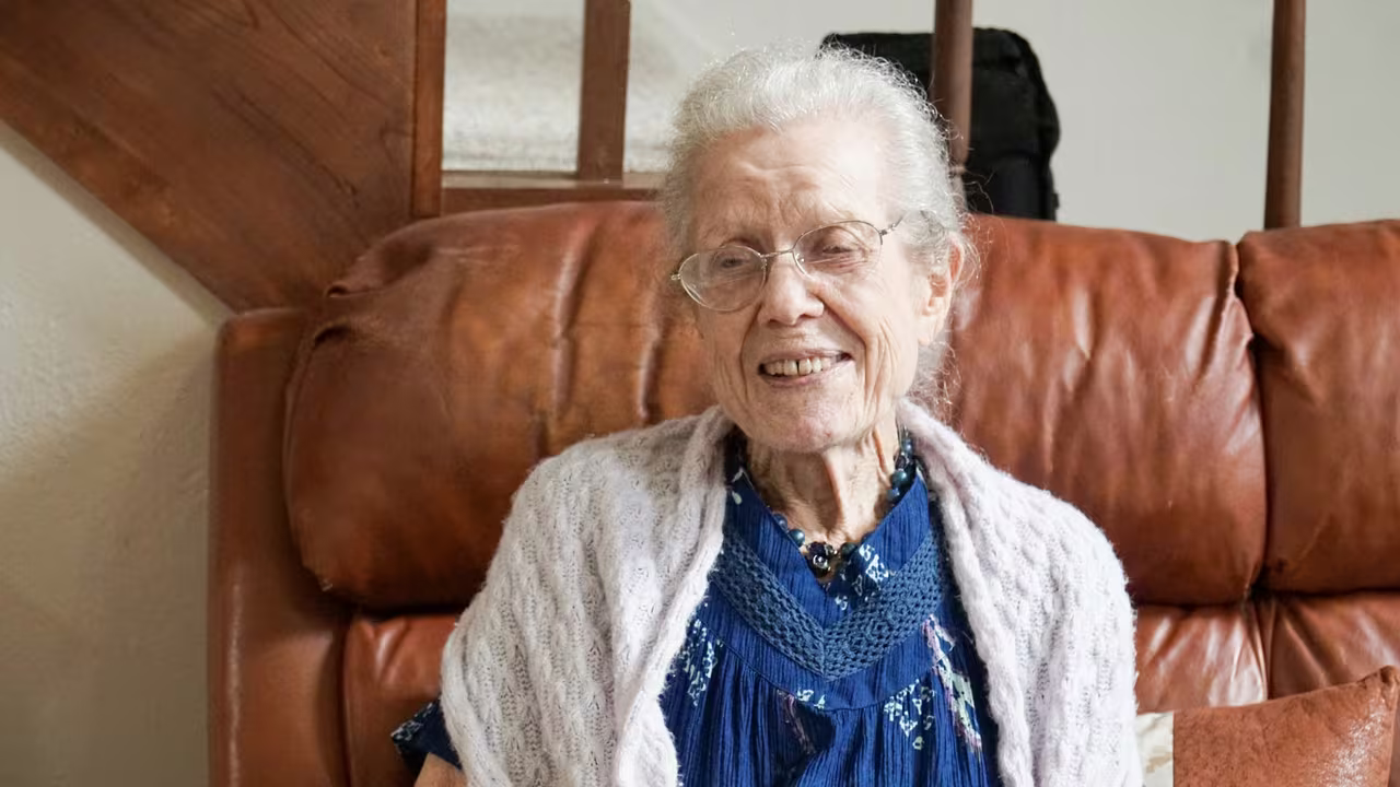 An older woman sitting smiling in an armchair at home, with a staircase behind her