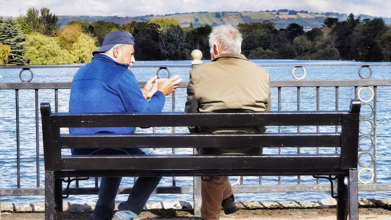 Two older men sitting together on a bench by a lake, talking